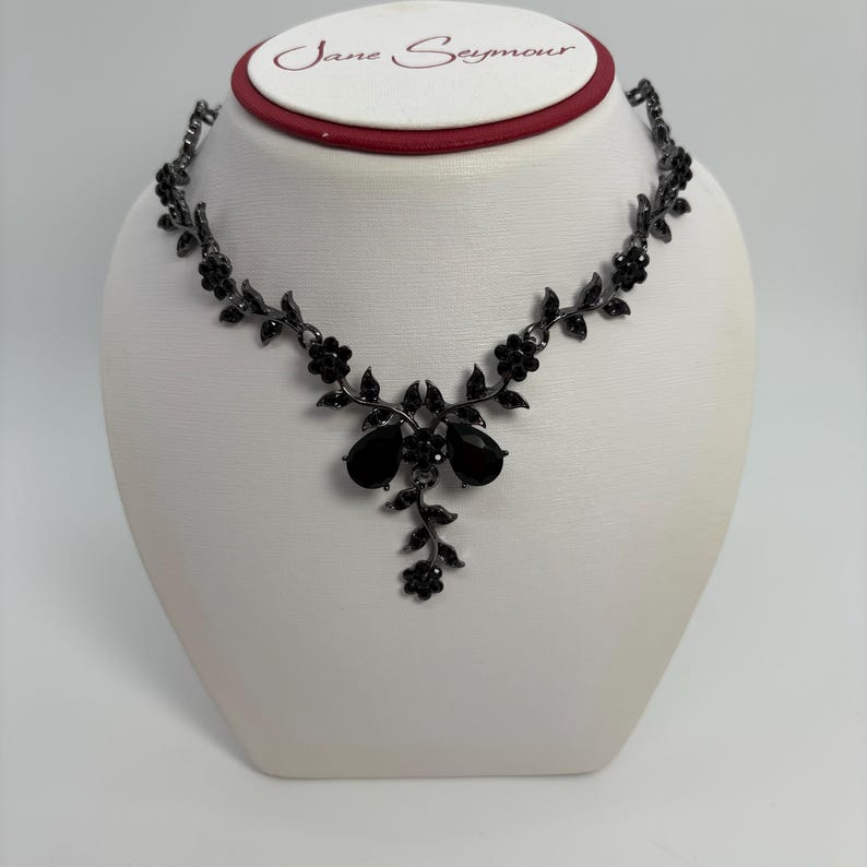 Pu&ograve; includere: Una collana floreale nera con pendenti a forma di goccia. La collana &egrave; esposta su un manichino bianco. Il marchio Jane Seymour &egrave; visibile nella parte superiore dell'immagine.