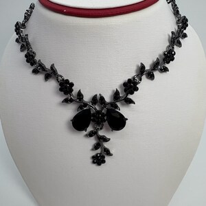 Pu&ograve; includere: Collana floreale nera con pendenti a goccia. La collana presenta un delicato design di fiori e foglie, con pietre nere. La collana &egrave; esposta su un manichino bianco.