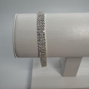 Könnte beinhalten: Ein silbernes Armband mit mehreren Reihen kleiner, funkelnder Strasssteine. Das Armband wird auf einem weißen, zylindrischen Schmuckhalter präsentiert, der sein Design und seine Textur hervorhebt. Das Schmuckstück ist für das Handgelenk konzipiert.