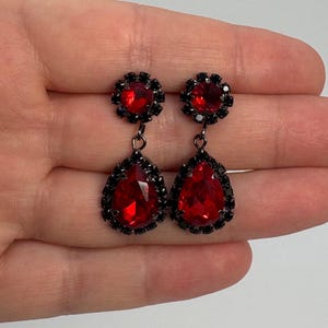 May include: A pair of drop earrings featuring a round red faceted stone at the top and a larger pear-shaped red faceted stone at the bottom. Each stone is surrounded by a border of small black crystals set in a dark metal frame with a link connector.