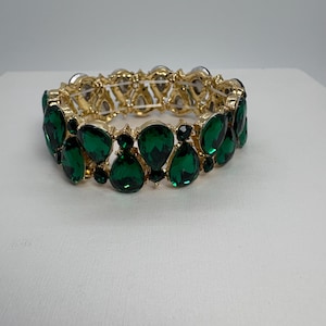 May include: A gold-toned bracelet featuring emerald green teardrop-shaped gemstones. The bracelet has a double-row design with the gemstones set in a gold metal frame. The gemstones are arranged in a repeating pattern.