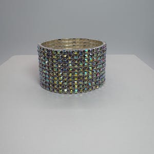 May include: A wide, iridescent rhinestone bracelet. The bracelet is made of multiple rows of small, colorful rhinestones set in a silver-tone metal. The stones reflect a rainbow of colors, creating a sparkling effect. This statement jewelry piece is perfect for special occasions.