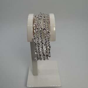 Puede incluir: Una pulsera plateada con múltiples hilos de brillantes pedrería. La pulsera se exhibe sobre un soporte blanco, resaltando su diseño. Los brillantes atrapan la luz, creando un efecto deslumbrante. La joya es adecuada para ocasiones especiales.