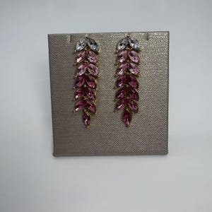 May include: A pair of dangling earrings with a leaf-like design. Each earring features a gold-colored metal frame adorned with marquise-cut crystals in shades of pink and clear. The earrings are displayed on a textured gray surface.