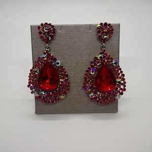 以下が含まれることがあります： 赤いしずく型のイヤリングのペア。各イヤリングは、小さな赤と虹色のクリスタルに囲まれた、大きなファセットカットの赤い宝石が特徴です。イヤリングは、灰色のスタンドに展示されています。