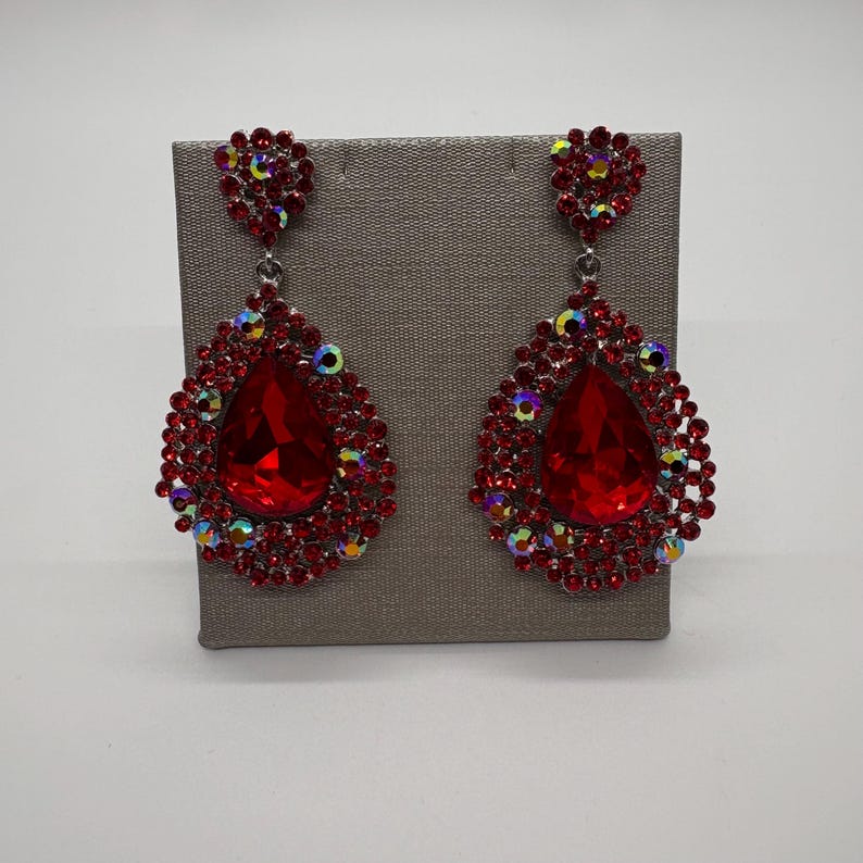 May include: A pair of red teardrop earrings. Each earring features a large, faceted red gem surrounded by smaller red and iridescent crystals. The earrings are displayed on a gray stand against a white background.