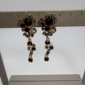 Op de afbeelding: Een paar goudkleurige bungelende oorbellen met donkerrode en amberkleurige strass-steentjes. De oorbellen hebben een wervelend bovendeel bezet met kleine kristallen en een bungelende onderketting van ronde stenen in verschillende maten.