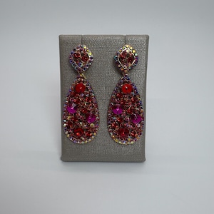 以下が含まれることがあります： 赤とピンクのきらびやかなティアドロップイヤリングのペア。各イヤリングは、赤とピンクのラインストーンのクラスターと、金色の正方形のトップが特徴です。イヤリングは、灰色のテクスチャード加工された表面に表示されています。