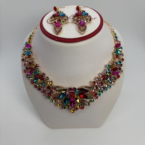 Op de afbeelding: Een goudkleurige ketting en bijpassende oorbellen set. De sieraden hebben een bloemmotief met een reeks kleurrijke, gefacetteerde edelstenen in tinten rood, blauw, groen, roze en geel. De oorbellen zijn van het druppeltype.