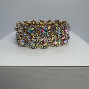 May include: Gold-toned stretch bracelet featuring iridescent, round, and oval-shaped rhinestones. The bracelet has a floral design with multiple rows of sparkling stones. The rhinestones reflect a rainbow of colors, including pink, blue, and green.