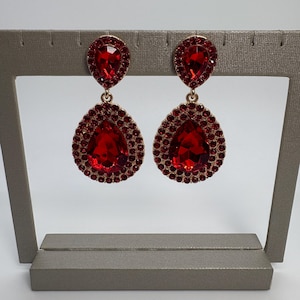 May include: A pair of drop earrings featuring a large pear-shaped red crystal center stone surrounded by a halo of smaller red rhinestones. Above the main drop, a smaller pear-shaped red crystal is set in a gold-toned metal frame.