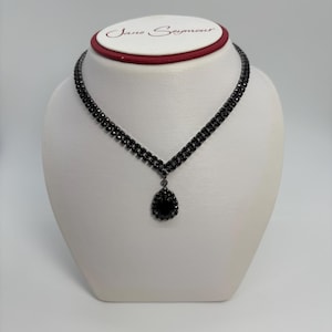 May include: A black necklace with a teardrop pendant is displayed on a white mannequin. The necklace has a double-strand design with small, dark stones. The pendant is a larger, teardrop-shaped stone. The brand name "Jane Seymour" is printed on the mannequin.