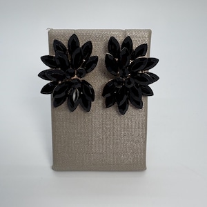 Puede incluir: Un par de pendientes de botón negros con forma floral. Cada pendiente presenta múltiples pétalos negros con forma de gema dispuestos alrededor de un grupo central. Los pendientes se exhiben sobre una superficie texturizada de tono neutro.