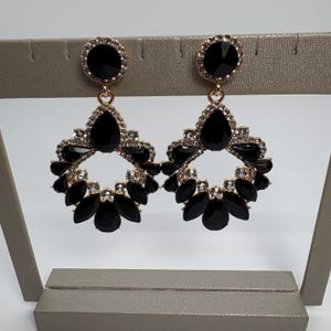 May include: A pair of gold-toned, statement earrings with black and clear gemstones. Each earring features a large, round black stone at the top, a teardrop-shaped black stone, and a floral-like design of black stones. Clear stones surround the black stones.