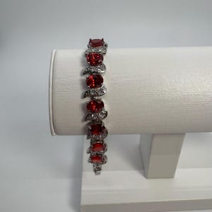 May include: A silver-toned bracelet featuring a series of oval-cut deep red stones alternating with small, clear, sparkling accent stones. The design consists of repeating links, each holding one central red stone surrounded by a curved metal frame.