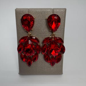 May include: A pair of dazzling red chandelier earrings. Each earring features a large, teardrop-shaped red gem at the top, connected to a cluster of smaller, similarly colored gems. The earrings have a gold-colored metal setting.