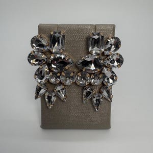 Puede incluir: Un par de pendientes dorados con cristales claros y facetados. Cada pendiente presenta cristales de diversas formas, como rectángulos, gotas y óvalos. Los cristales están engarzados sobre una base dorada, creando un diseño glamuroso y llamativo.