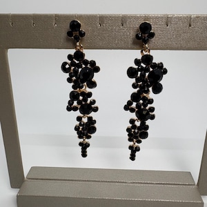 Puede incluir: Un par de pendientes colgantes con un diseño de racimo. Los pendientes presentan piedras preciosas negras facetadas y metal dorado. Las piedras preciosas están dispuestas en un patrón en cascada, creando un accesorio glamuroso y llamativo.