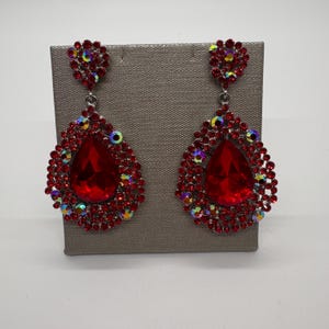 May include: A pair of red teardrop earrings with a cluster of small, iridescent rhinestones at the top and around the large, faceted red center stone. The earrings are displayed on a gray surface.