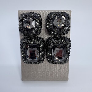 Puede incluir: Cuatro pendientes cuadrados de color gris oscuro con grandes piedras centrales facetadas. Cada pendiente está rodeado de cristales más pequeños a juego, creando un borde brillante y texturizado. Los pendientes se exhiben sobre una superficie gris claro.