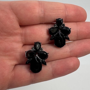 Könnte beinhalten: Ein Paar Ohrstecker mit Clustern aus facettierten schwarzen Steinen in Tropfen- und Ovalform. Die Steine sind in einen dunklen Metallrahmen gefasst und bilden ein symmetrisches, facettiertes Design, das das Licht auf den dunklen Oberflächen reflektiert.