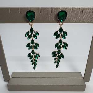 May include: A pair of dangling earrings with a gold-tone metal setting. Each earring features a teardrop-shaped green gem at the top, with a cascading design of green leaf-shaped gems. The earrings are displayed on a gray jewelry stand.