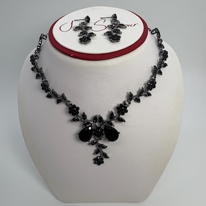 Pu&ograve; includere: Un set di collana e orecchini neri con un design floreale. La collana presenta un motivo a forma di vite con fiori neri e due grandi pietre nere a forma di goccia. Gli orecchini riprendono il design floreale. I gioielli sono esposti su un busto bianco.
