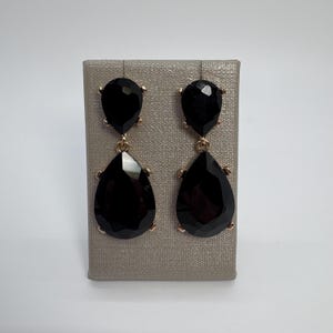 Puede incluir: Un par de pendientes negros en forma de lágrima con detalles dorados. Los pendientes se exhiben sobre una superficie gris texturizada. Los pendientes tienen un poste dorado y un marco dorado alrededor de las piedras negras en forma de lágrima.