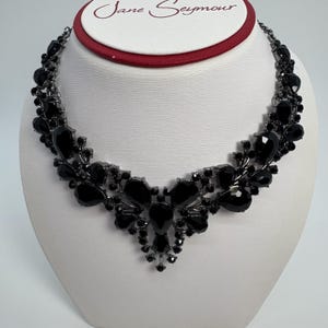 以下が含まれることがあります： 黒いステートメントネックレス、フローラルデザイン。 複数の涙型と丸い黒い宝石が、暗い金属フレームにセットされています。 ネックレスは、白いバストに表示され、上部に「Jane Seymour」の文字があります。