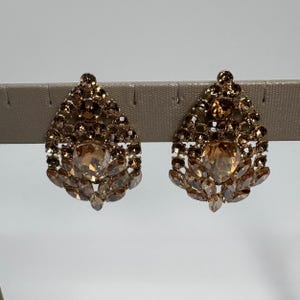 May include: A pair of teardrop-shaped earrings featuring a cluster of light brown and champagne-colored faceted crystals set in a metallic frame. The design includes a large central stone surrounded by smaller round and marquise-cut crystals.