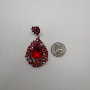 May include: A large, red teardrop-shaped earring with a cluster of smaller red and iridescent stones. The earring is silver-toned and has a smaller floral-shaped cluster at the top. A quarter is next to the earring for size comparison.