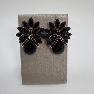 May include: A pair of black statement earrings with a floral design. Each earring features a cluster of black, teardrop-shaped gems and smaller, round black gems set in a gold-tone metal. The earrings are displayed on a neutral-toned surface.