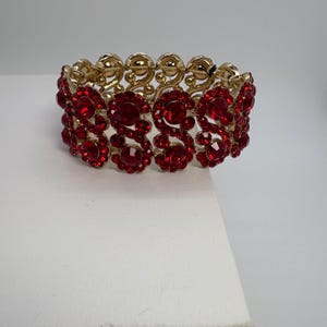 Puede incluir: Una pulsera dorada con piedras preciosas rojas vibrantes. La pulsera presenta un diseño elástico con secciones alternas de eslabones dorados ornamentados y grupos de joyas rojas redondas. Las piedras preciosas están muy juntas, creando un accesorio llamativo.