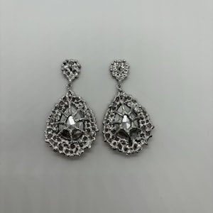 May include: A pair of silver-toned drop earrings. Each earring features a textured, openwork design surrounding a central, faceted, clear gem. The earrings have a teardrop shape and a small, decorative top.