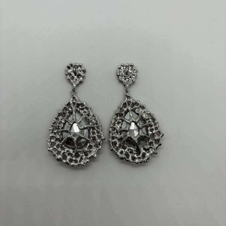 May include: A pair of silver-toned drop earrings. Each earring features a textured, openwork design surrounding a central, faceted, clear gem. The earrings have a teardrop shape and a small, decorative top.