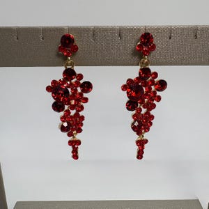 May include: A pair of gold-tone drop earrings featuring a cluster of vibrant red faceted crystals. Each earring has a single round crystal at the top, connected to a larger, asymmetrical arrangement of various sized red stones that taper downward.