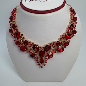 Może przedstawiać: Złoty naszyjnik z dużymi, fasetowanymi czerwonymi kamieniami szlachetnymi. Naszyjnik jest prezentowany na białym popiersiu, podkreślając jego skomplikowany design i żywy kolor. Kamienie szlachetne ułożone są we wzór kwiatowy.