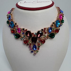Op de afbeelding: Een goudkleurige statementketting met een reeks kleurrijke, facetgeslepen edelstenen. De ketting toont een verscheidenheid aan juweeltinten, waaronder rood, roze, blauw, groen en paars. De ketting wordt getoond op een witte buste.