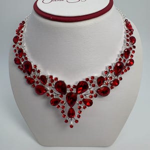 Może przedstawiać: Srebrny naszyjnik z kaskadą dużych, czerwonych kamieni w kształcie łez. Mniejsze czerwone kamienie zdobią wzór, tworząc błyszczący, elegancki wygląd. Naszyjnik jest prezentowany na białym popiersiu, ukazując jego misterne detale.