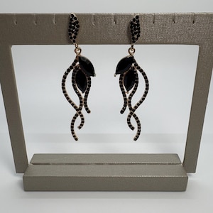 Puede incluir: Un par de pendientes colgantes negros y dorados. Cada pendiente presenta una gema negra en forma de hoja y una serie de líneas doradas curvas adornadas con pequeñas gemas negras. Los pendientes se exhiben en un expositor de joyería gris.