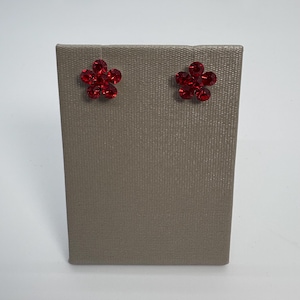 May include: Two red flower-shaped stud earrings on a gray display stand. The earrings are made of small, sparkling red stones. The display stand is rectangular and has a textured surface.