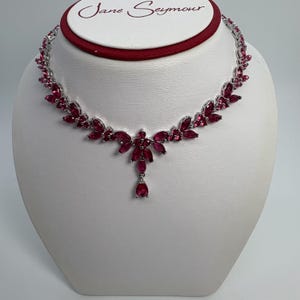 May include: A silver-toned necklace with a floral design, featuring numerous marquise-cut, ruby-colored gemstones. The necklace is displayed on a white bust, with the brand name "Jane Seymour" visible at the top.