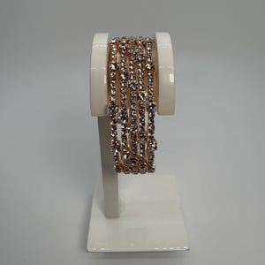 Puede incluir: Un conjunto de brazaletes de oro rosa con brillantes pedrería transparente. Los brazaletes se exhiben en un soporte blanco para joyas, mostrando su diseño delicado y sus intrincados detalles. La joyería es perfecta para añadir un toque de elegancia a cualquier atuendo.