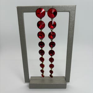 May include: A pair of long, dangling earrings featuring graduated, round, faceted red crystals. The crystals are connected by gold-toned metal links, creating a cascading effect. The earrings are displayed on a gray stand against a white background.