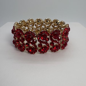 May include: A gold-toned stretch bracelet adorned with vibrant red gemstones. The bracelet features a repeating floral design with the red stones set in a gold metal frame. This piece of jewelry is suitable for special occasions.