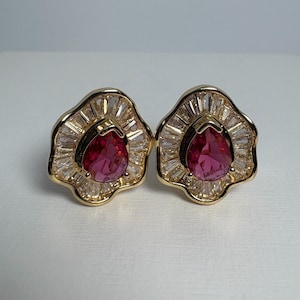 May include: A pair of gold-coloured stud earrings. Each earring has a teardrop-shaped, deep red gemstone at the centre, surrounded by a halo of clear, baguette-cut stones. The earrings have a floral-like design.