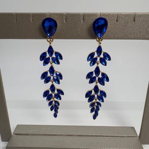 May include: A pair of dangling earrings with a gold-tone metal setting. Each earring features a large, teardrop-shaped blue gemstone at the top, with a cascading design of smaller, leaf-shaped blue gemstones. The earrings are displayed on a neutral-colored stand.