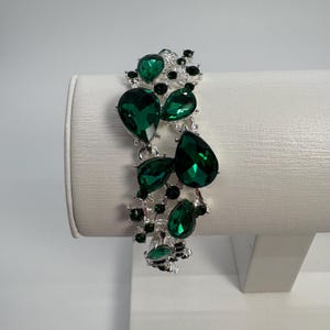 May include: A silver-toned bracelet featuring large, emerald-green teardrop-shaped gemstones and smaller, round green stones. The bracelet is displayed on a white stand, showcasing its intricate design and sparkling appearance.