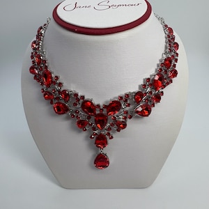 Puede incluir: Un collar plateado con una cascada de vibrantes piedras preciosas rojas. El collar presenta un colgante central y un diseño simétrico, exhibido sobre un busto blanco. El nombre de la marca "Jane Seymour" es visible en la parte superior.