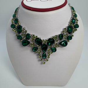 Puede incluir: Un collar plateado con un diseño floral. El collar presenta grandes piedras en forma de lágrima de color verde esmeralda y piedras más pequeñas de color verde claro. El collar se exhibe en un busto blanco.